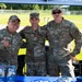 Three U.S. Air Force personnel in OCP (Operational Camouflage Pattern) uniforms pose together at an outdoor recruitment or outreach booth. The table in front of them displays Air Force Reserve and U.S. Air Force recruitment brochures and materials. The event appears to be taking place at an outdoor venue with a green field visible in the background.