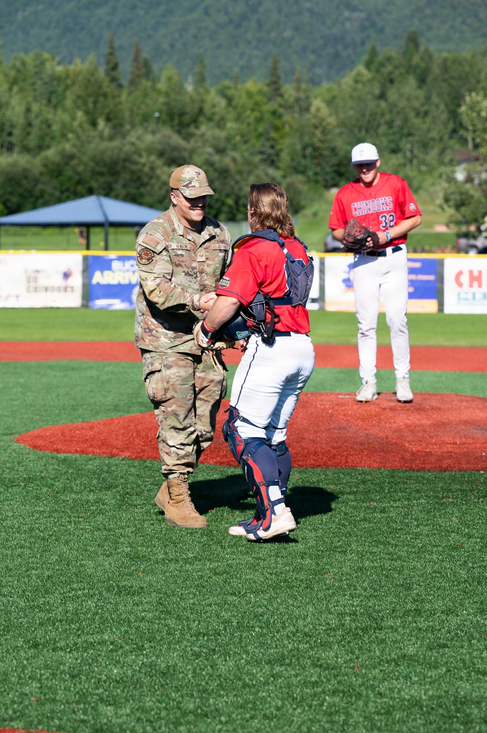 A military service member in OCP (Operational Camouflage Pattern) uniform is shaking hands with a baseball catcher on the pitcher's mound, likely after throwing a ceremonial first pitch. A pitcher wearing jersey number 32 for the 'Chinooks' team stands in the background on the mound. The event appears to take place at an outdoor baseball stadium with green mountains and trees visible in the background.