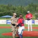 A military service member in OCP (Operational Camouflage Pattern) uniform is shaking hands with a baseball catcher on the pitcher's mound, likely after throwing a ceremonial first pitch. A pitcher wearing jersey number 32 for the 'Chinooks' team stands in the background on the mound. The event appears to take place at an outdoor baseball stadium with green mountains and trees visible in the background.