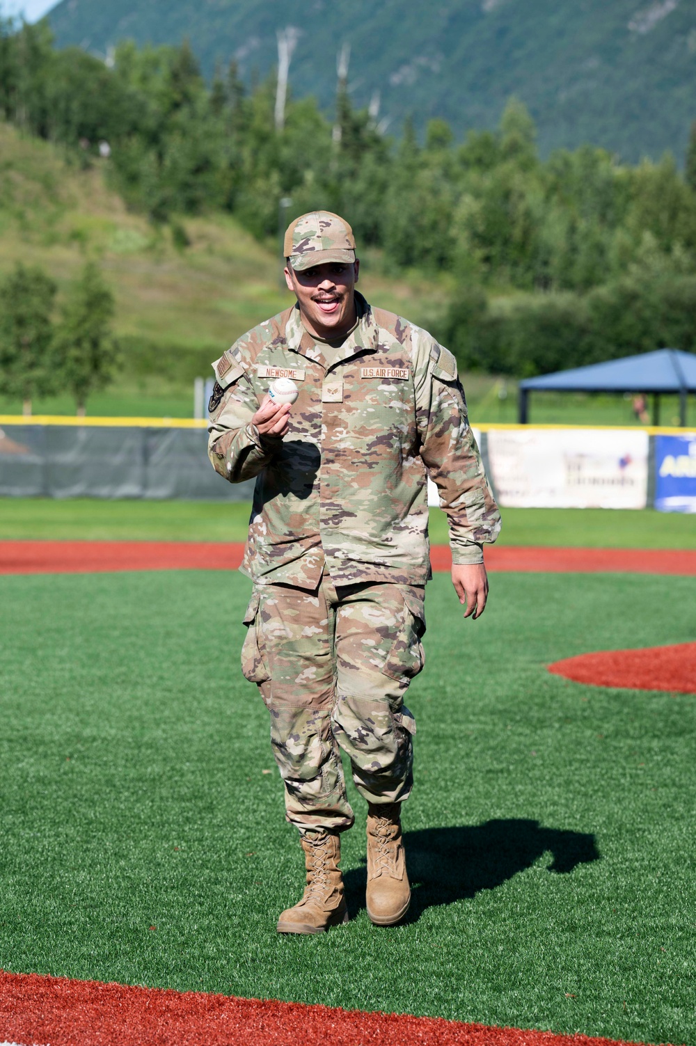 A U.S. Air Force service member wearing OCP (Operational Camouflage Pattern) uniform with the name tape 'NEWSOME' is walking on a baseball field while holding a baseball. The individual appears to be smiling and in good spirits, likely having just thrown or preparing to throw a ceremonial first pitch. The setting is an outdoor baseball stadium with green turf, red warning track, outfield wall with advertisements, and a mountainous, tree-covered backdrop.