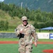 A U.S. Air Force service member wearing OCP (Operational Camouflage Pattern) uniform with the name tape 'NEWSOME' is walking on a baseball field while holding a baseball. The individual appears to be smiling and in good spirits, likely having just thrown or preparing to throw a ceremonial first pitch. The setting is an outdoor baseball stadium with green turf, red warning track, outfield wall with advertisements, and a mountainous, tree-covered backdrop.