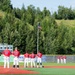A baseball game is taking place at Lee Jordan Field, with players from two teams lined up along the outfield grass facing an American flag on a flagpole, likely during the national anthem. The home team wears red uniforms while the opposing team wears white. The field is surrounded by lush green trees and hills under a bright, sunny sky.