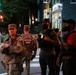 The image shows National Guard personnel standing at a street corner at night alongside several civilians. Two soldiers in camouflage uniforms with 'National Guard' patches are visible, positioned near a crosswalk. The setting appears to be an urban intersection with traffic signals and city buildings in the background.