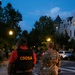 The image shows two officials walking across a crosswalk at dusk in what appears to be an urban residential neighborhood. One individual is wearing a black tactical vest labeled 'CSOSA' (Court Services and Offender Supervision Agency) in orange lettering, while the other is in full military camouflage uniform with a 'NATIONAL GUARD' patch on their back. The setting appears to be Washington, D.C., based on the Victorian-style architecture and agency identification.