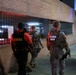 A nighttime scene showing National Guard soldiers and a CSOSA (Court Services and Offender Supervision Agency) officer walking along a sidewalk next to a brick building with iron security fencing. The group appears to be conducting a patrol or joint operation in an urban area. A civilian in a white shirt is also visible on the left side of the frame.