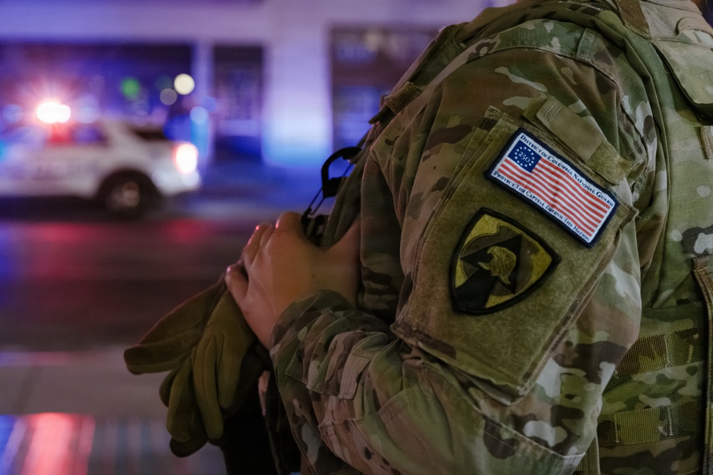 A close-up photograph of a military service member in MultiCam (OCP) camouflage uniform, showing the right shoulder sleeve with two distinct patches. The image was taken at night with emergency vehicle lights visible in the blurred background. The soldier appears to be holding or carrying equipment.