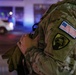 A close-up photograph of a military service member in MultiCam (OCP) camouflage uniform, showing the right shoulder sleeve with two distinct patches. The image was taken at night with emergency vehicle lights visible in the blurred background. The soldier appears to be holding or carrying equipment.