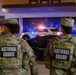 Two National Guard personnel are standing on a street at night, facing away from the camera toward a police vehicle with active emergency lights. Both individuals are wearing camouflage uniforms with 'NATIONAL GUARD' vests and military caps with name tapes visible. The scene suggests a law enforcement or civil support operation in an urban environment.