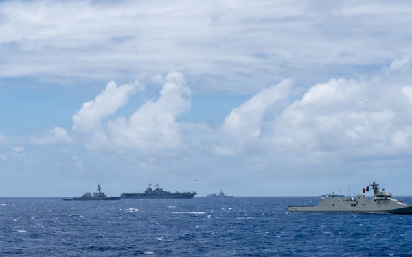 A multinational naval formation is underway on open ocean, with multiple warships from different nations sailing in loose formation. The scene appears to depict a joint naval exercise or fleet review, with vessels spread across the horizon. At least one helicopter is visible in the air above the formation.