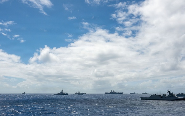 A large multinational naval formation is underway across open ocean waters, with numerous warships arranged in a broad line formation stretching across the horizon. The vessels appear to be conducting a coordinated fleet exercise or passage, consistent with a multinational naval exercise. The image is taken from the deck or superstructure of another vessel participating in the formation.