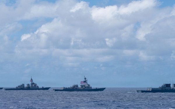 The image shows a formation of four naval warships sailing in open ocean under a partly cloudy sky. At least two national flags are visible — an American flag and what appears to be a South Korean flag — suggesting a multinational naval exercise or patrol. The ships appear to be destroyers or guided-missile destroyers operating in close formation.