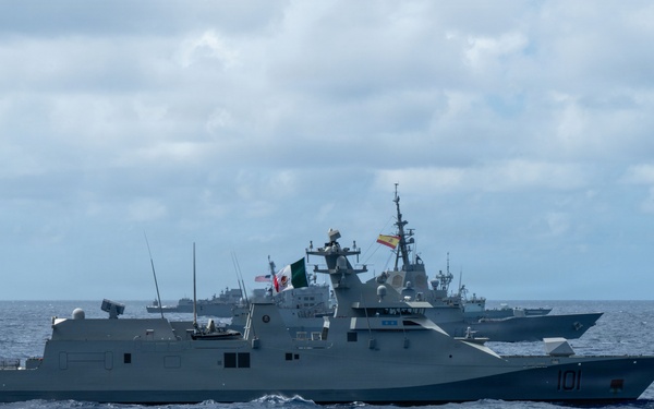 The image shows a formation of multiple naval warships underway at sea under partly cloudy skies. The lead vessel, numbered '101', is flying what appears to be a Mexican naval flag, while a second vessel behind it displays what appears to be a Spanish flag. At least three to four warships are visible in the formation, suggesting a multinational naval exercise or transit.