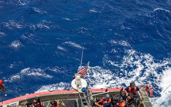 An aerial photograph shows a U.S. Coast Guard rigid-hull inflatable boat (RHIB) numbered 26281 operating in open blue ocean waters. The vessel carries multiple personnel in tactical gear and life vests, with an American flag visible at the stern. The boat appears to be either being deployed or recovered, as tethering lines are visible extending from both sides.