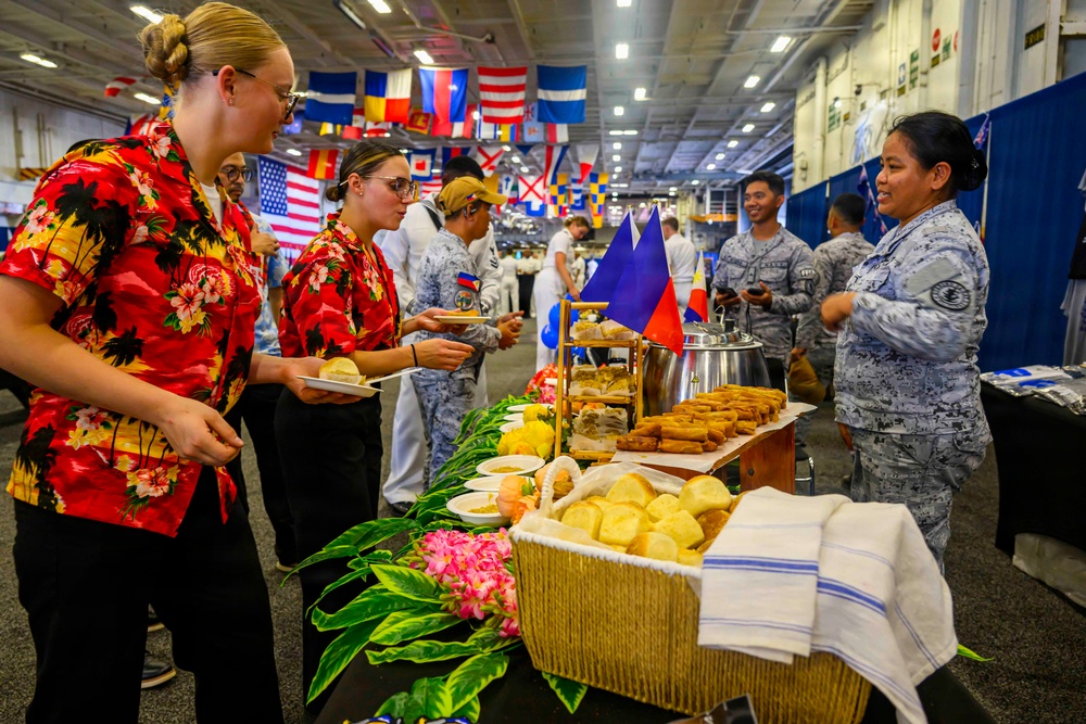 A multinational military cultural food fair or international day event is taking place in what appears to be a large ship hangar deck or military facility. Personnel in various uniforms and civilian attire are sampling food from decorated tables representing different nations, with international flags displayed overhead. A Philippine flag is prominently visible at one food station, and two individuals in matching Hawaiian shirts appear to be serving or hosting.