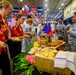 A multinational military cultural food fair or international day event is taking place in what appears to be a large ship hangar deck or military facility. Personnel in various uniforms and civilian attire are sampling food from decorated tables representing different nations, with international flags displayed overhead. A Philippine flag is prominently visible at one food station, and two individuals in matching Hawaiian shirts appear to be serving or hosting.