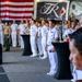 A formal military ceremony or reception is taking place in what appears to be a ship's hangar bay, likely aboard USS Theodore Roosevelt (indicated by 'The Big Stick' banner). Personnel in U.S. Navy white dress uniforms, U.S. Marine Corps uniforms, and what appear to be Japanese Maritime Self-Defense Force uniforms are standing at attention or in formation. The presence of both American and Japanese flags suggests a bilateral military event or joint ceremony.