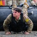 A soldier in MultiCam/OCP camouflage uniform is performing a push-up or low crawl exercise on a gravel surface beneath a large black inflatable rubber boat (IRB/Zodiac). The soldier wears a black plate carrier/vest and displays a yellow identification tag marked 'A5' on the left sleeve, along with a US flag patch, indicating a military training or selection event. Water droplets are visible in the air, suggesting wet/rainy conditions or recent water immersion.