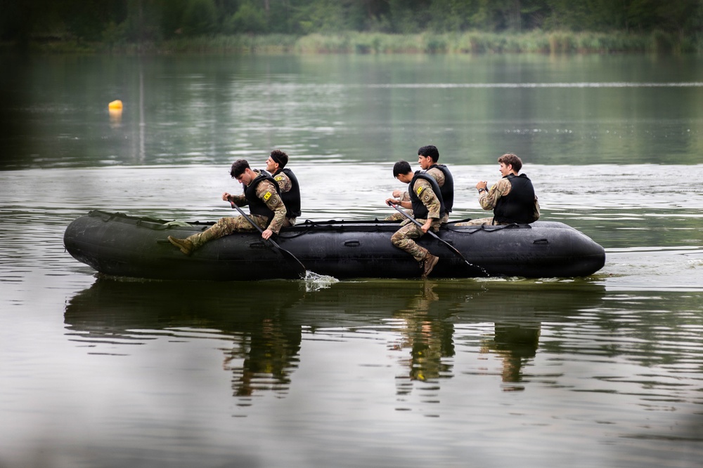 Five individuals in military camouflage uniforms and black life vests are paddling a black inflatable rubber assault boat (zodiac-style) across a calm lake or river. The personnel appear to be engaged in a military training exercise, sitting along the sides of the craft and using paddles. The background features dense green treeline and overcast lighting conditions.