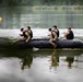 Five individuals in military camouflage uniforms and black life vests are paddling a black inflatable rubber assault boat (zodiac-style) across a calm lake or river. The personnel appear to be engaged in a military training exercise, sitting along the sides of the craft and using paddles. The background features dense green treeline and overcast lighting conditions.
