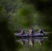 Three individuals in military camouflage uniforms and black tactical vests are paddling a black inflatable rubber boat across a calm, reflective body of water. The scene is framed by out-of-focus green foliage in the foreground, suggesting the photo was taken from a concealed or distant vantage point. The lush green tree line in the background indicates a temperate, forested environment.