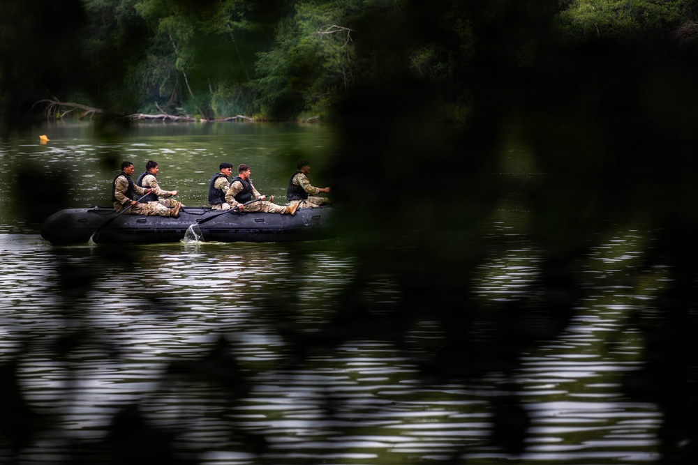 A group of approximately five individuals in military camouflage uniforms and tactical vests are navigating a black inflatable rubber boat across a calm river. The image is captured through partially obscuring foreground foliage, giving a surveillance-like perspective. The background features dense green tree cover along the riverbank.