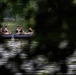 A group of approximately five individuals in military camouflage uniforms and tactical vests are navigating a black inflatable rubber boat across a calm river. The image is captured through partially obscuring foreground foliage, giving a surveillance-like perspective. The background features dense green tree cover along the riverbank.