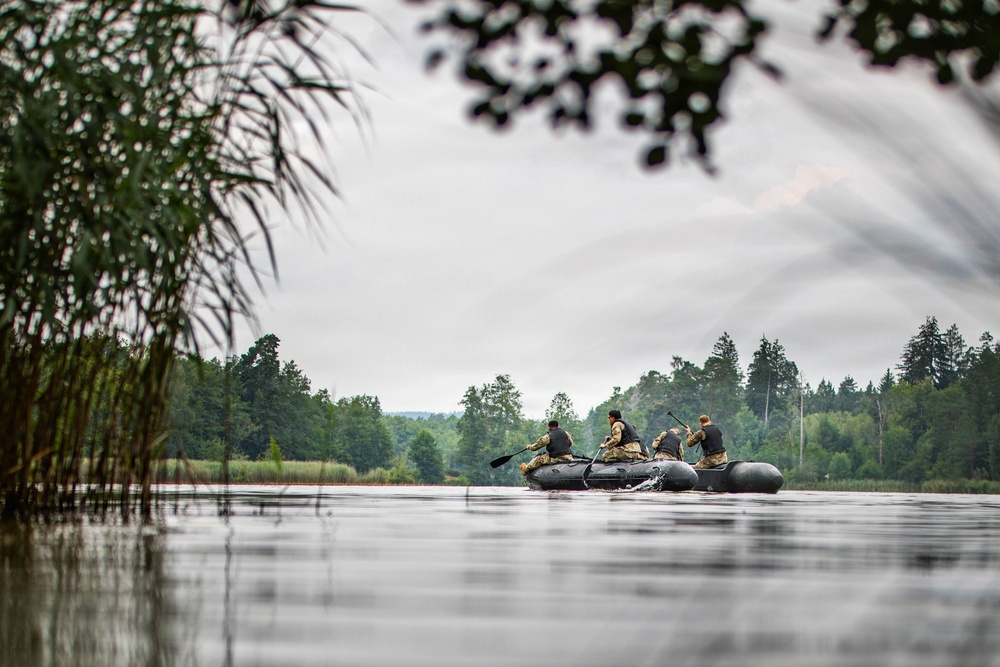 Several individuals wearing camouflage and dark tactical clothing are paddling two dark inflatable rubber boats across a calm lake or river. The scene is framed by reeds and tree branches in the foreground, with a dense treeline visible in the background under an overcast sky. The composition suggests a military training exercise or tactical water crossing operation.