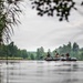 Several individuals wearing camouflage and dark tactical clothing are paddling two dark inflatable rubber boats across a calm lake or river. The scene is framed by reeds and tree branches in the foreground, with a dense treeline visible in the background under an overcast sky. The composition suggests a military training exercise or tactical water crossing operation.