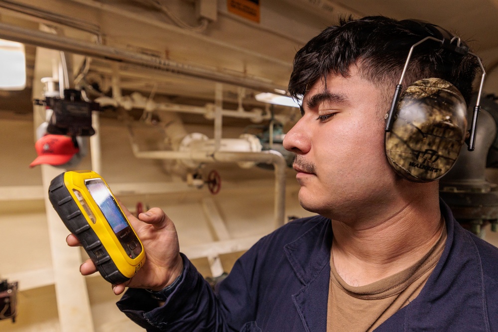 A young man wearing camouflage-pattern ear protection/hearing muffs is closely examining a rugged yellow handheld device with a touchscreen display. He appears to be in an industrial or mechanical space, likely aboard a ship or in a facility with visible pipes, valves, and conduits in the background. He is wearing a dark navy work shirt over a brown undershirt.