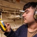 A young man wearing camouflage-pattern ear protection/hearing muffs is closely examining a rugged yellow handheld device with a touchscreen display. He appears to be in an industrial or mechanical space, likely aboard a ship or in a facility with visible pipes, valves, and conduits in the background. He is wearing a dark navy work shirt over a brown undershirt.