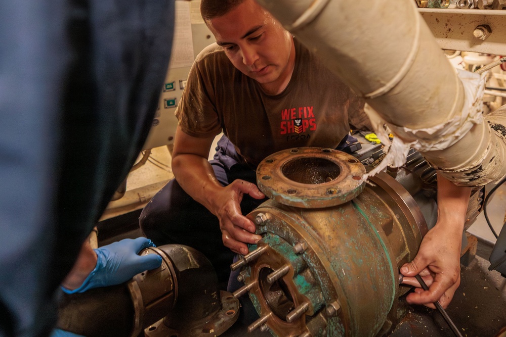 A technician wearing a brown 'WE FIX SHIPS' t-shirt is working on a large corroded industrial pump or valve assembly, assisted by at least two other individuals. The equipment shows significant oxidation and wear, suggesting it is part of a maritime or naval vessel's mechanical system. The work appears to involve disassembly or maintenance of a flanged pipe fitting.
