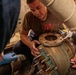 A technician wearing a brown 'WE FIX SHIPS' t-shirt is working on a large corroded industrial pump or valve assembly, assisted by at least two other individuals. The equipment shows significant oxidation and wear, suggesting it is part of a maritime or naval vessel's mechanical system. The work appears to involve disassembly or maintenance of a flanged pipe fitting.