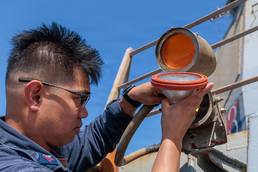 A young man wearing glasses and a dark uniform is performing maintenance on a nautical signal light or navigation lamp mounted on what appears to be a vessel or maritime structure. He is carefully handling the lamp assembly, which has an orange/amber lens cover that is open, revealing the white reflector bulb housing beneath. The work is being conducted outdoors under a clear blue sky.