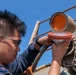 A young man wearing glasses and a dark uniform is performing maintenance on a nautical signal light or navigation lamp mounted on what appears to be a vessel or maritime structure. He is carefully handling the lamp assembly, which has an orange/amber lens cover that is open, revealing the white reflector bulb housing beneath. The work is being conducted outdoors under a clear blue sky.