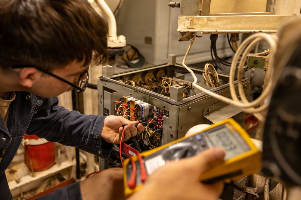 A technician wearing glasses and a blue shirt is using probe leads to test an open electrical control panel or equipment cabinet filled with wiring, relays, and components. A second person is holding a yellow Fluke digital multimeter, reading measurements from the display. The setting appears to be an industrial or mechanical room with aging equipment.