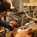 A technician wearing glasses and a blue shirt is using probe leads to test an open electrical control panel or equipment cabinet filled with wiring, relays, and components. A second person is holding a yellow Fluke digital multimeter, reading measurements from the display. The setting appears to be an industrial or mechanical room with aging equipment.