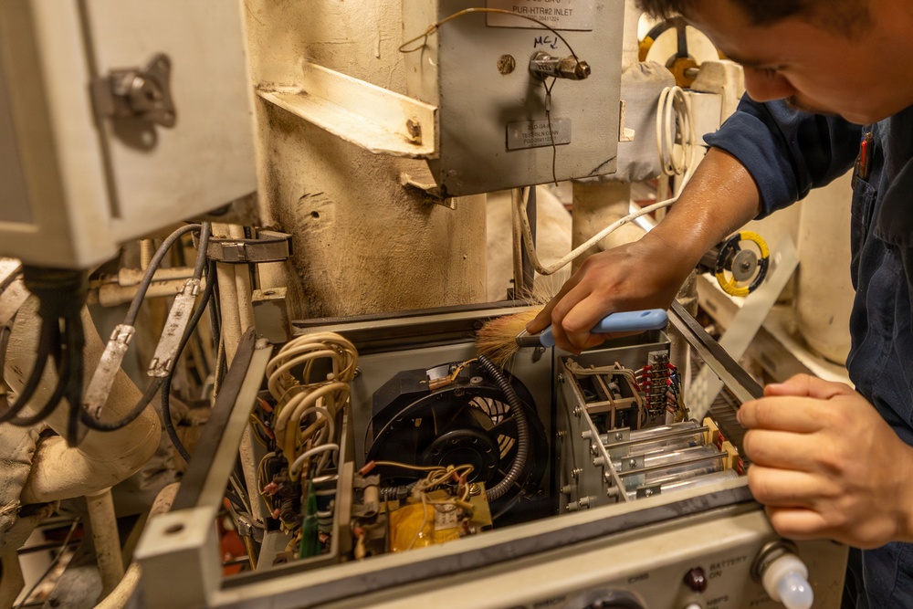 A technician in a dark blue uniform is performing maintenance on an open electronic equipment panel, using a natural-bristle brush to clean internal components. The equipment appears to be aged industrial or military-grade machinery with visible wiring, a cooling fan, and circuit components. The setting suggests a confined mechanical or engineering space, possibly aboard a naval vessel or industrial facility.