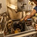 A technician in a dark blue uniform is performing maintenance on an open electronic equipment panel, using a natural-bristle brush to clean internal components. The equipment appears to be aged industrial or military-grade machinery with visible wiring, a cooling fan, and circuit components. The setting suggests a confined mechanical or engineering space, possibly aboard a naval vessel or industrial facility.