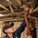 A Navy sailor in dark blue coveralls uses a large heavy-duty pipe wrench to work on overhead piping in what appears to be a shipboard engineering or mechanical space. A second sailor in the background observes the work. The environment is consistent with the interior of a U.S. Navy vessel, featuring dense overhead pipe runs, cable bundles, and painted metal bulkheads.