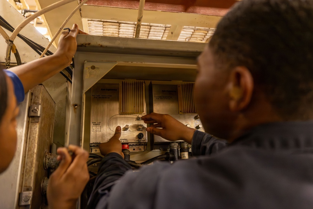 Two technicians are working on an open electrical or mechanical control panel, appearing to inspect or service internal components. The panel contains what appear to be voltage regulators, heat sinks, and various control components. The setting appears to be an industrial or military/naval environment based on the equipment style and construction.