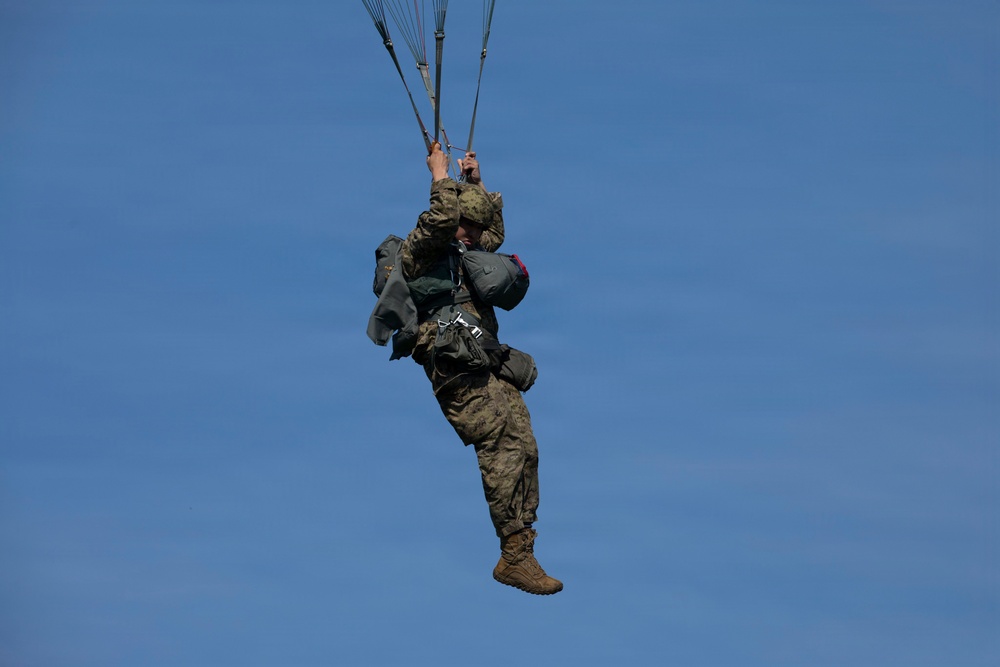 A military paratrooper is descending under a parachute canopy against a clear blue sky. The individual is wearing full combat gear including camouflage uniform, helmet, and multiple equipment packs. The parachute canopy is not fully visible in the frame, but the suspension lines are clearly seen converging above the jumper.