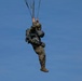 A military paratrooper is descending under a parachute canopy against a clear blue sky. The individual is wearing full combat gear including camouflage uniform, helmet, and multiple equipment packs. The parachute canopy is not fully visible in the frame, but the suspension lines are clearly seen converging above the jumper.