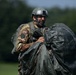 A military paratrooper wearing camouflage uniform and a tactical helmet numbered '22' is gathering and carrying a deployed parachute after landing on a grassy field. The soldier appears to be in a post-jump recovery phase, bundling the dark-colored parachute canopy and lines in his arms. The background shows a green, tree-lined open field under clear sky conditions.