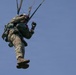 A military paratrooper is suspended in mid-air against a clear blue sky, captured from a low angle looking upward. The individual is wearing full military combat gear including a camouflage uniform, helmet with goggles, tactical gloves, and a parachute harness system. The parachute lines are visible extending upward out of frame.