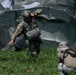 A military paratrooper has just landed on a grassy field and is managing their deployed parachute canopy. The soldier is wearing full combat gear including a helmet, multicam uniform, and harness with attached equipment pack. A second individual in lighter-colored gear is visible in the foreground, out of focus.