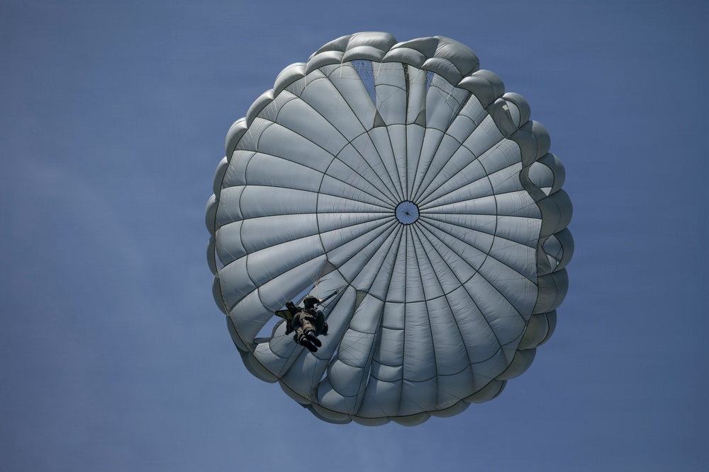 A military-style parachutist is descending beneath a large, round gray/silver parachute canopy against a clear blue sky. The parachute appears to be a round military static-line type, fully deployed and inflated. The jumper is visible suspended below the canopy in full gear.