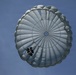A military-style parachutist is descending beneath a large, round gray/silver parachute canopy against a clear blue sky. The parachute appears to be a round military static-line type, fully deployed and inflated. The jumper is visible suspended below the canopy in full gear.