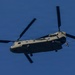 A United States Army CH-47 Chinook tandem-rotor helicopter is photographed in flight against a clear blue sky. The aircraft is viewed from below at a slight angle, showing its distinctive dual-rotor configuration. Personnel appear to be rappelling or fast-roping from the rear of the aircraft.