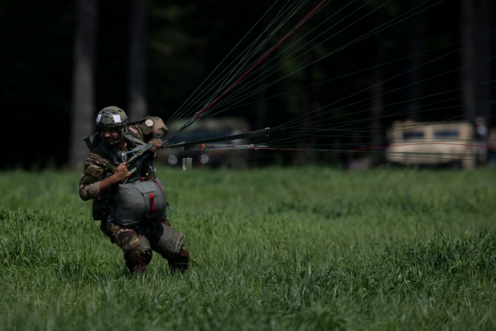 A military paratrooper has just landed in a grassy field and is managing their parachute lines, which extend behind them. The soldier is wearing full combat gear including a helmet, camouflage uniform, and a large pack. Military vehicles are visible in the blurred background near a tree line.