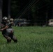 A military paratrooper has just landed in a grassy field and is managing their parachute lines, which extend behind them. The soldier is wearing full combat gear including a helmet, camouflage uniform, and a large pack. Military vehicles are visible in the blurred background near a tree line.